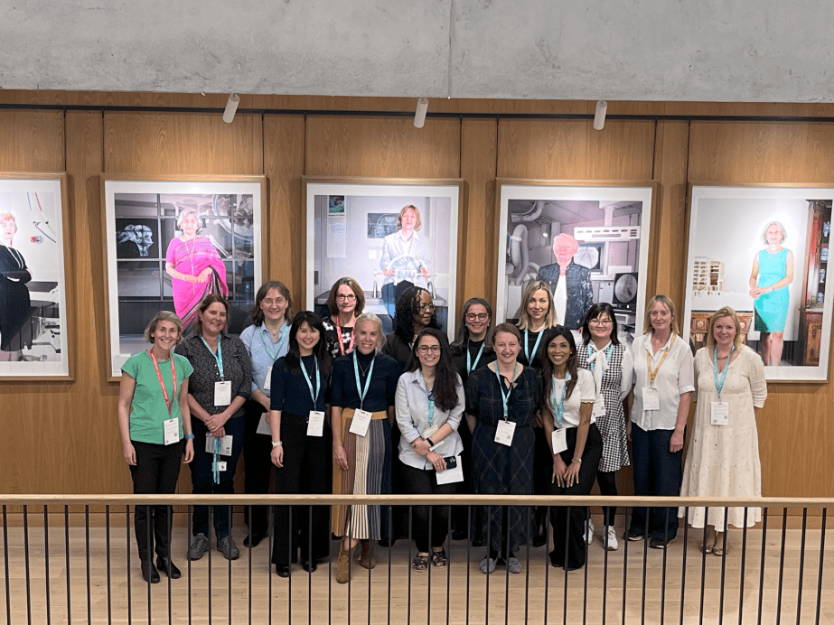Image of a group of people smiling. They are wearing lanyards and name tags. There are portraits of Women in Surgery behind them.